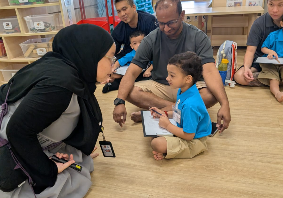 Teacher kneels to speak with a seated child holding a clipboard, with parents and kids on the floor behind.